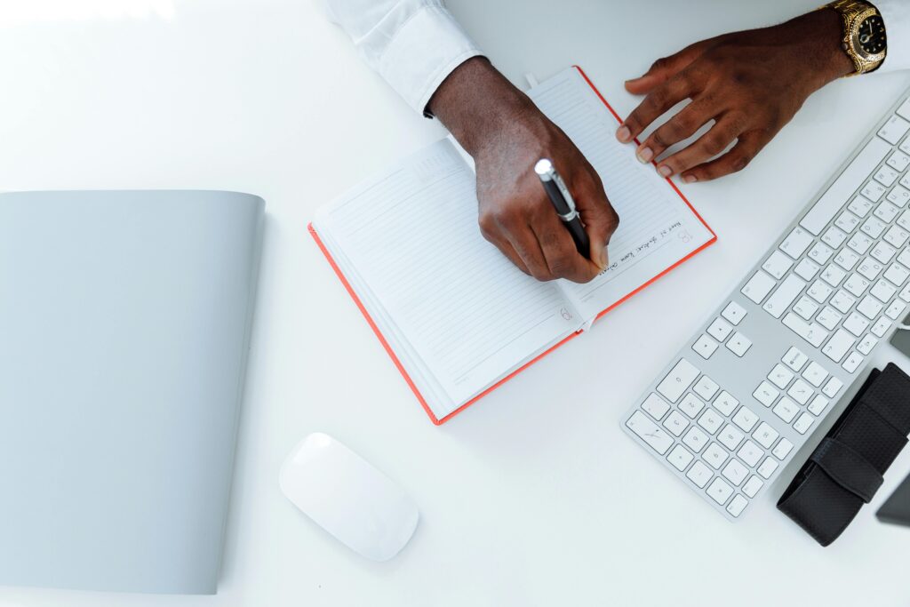 pexels-photo-5060557-5060557 Businessman writing in a notebook on a white desk with keyboard and mouse.